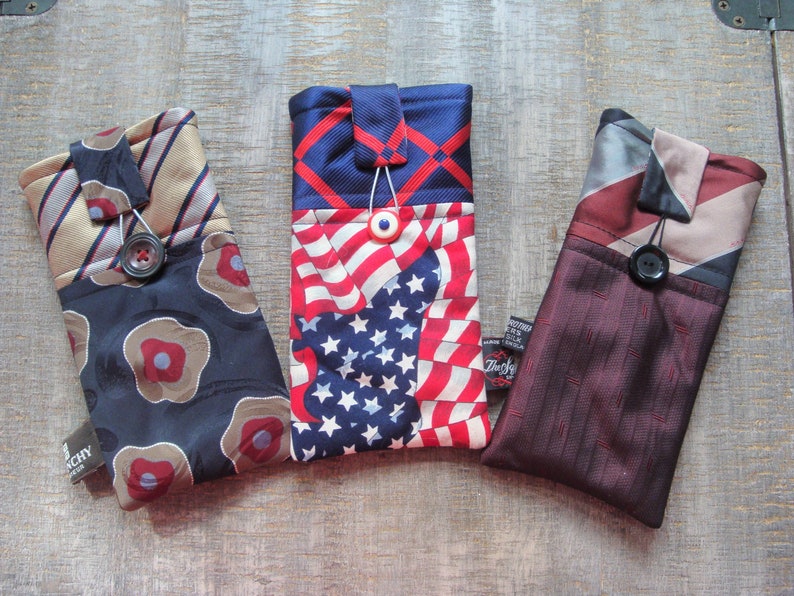 May include: Three handmade phone cases crafted from repurposed neckties. The cases feature various patterns, including stripes, floral designs, and an American flag motif. Each case has a button closure and a small tag.