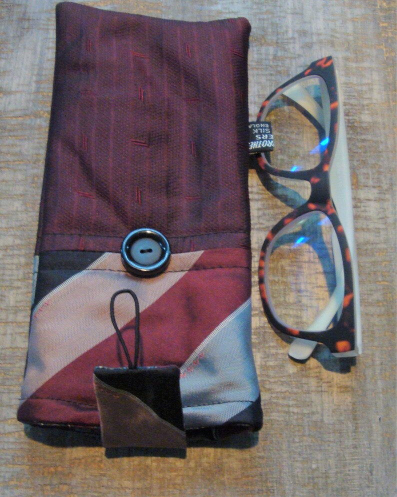 May include: A handmade glasses case and a pair of eyeglasses. The case is made of burgundy and striped fabric with a black button closure. The eyeglasses have a tortoiseshell pattern frame. A small, square, leather-like tag hangs from the case.