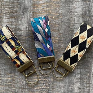 May include: Three fabric key fobs with bronze-colored hardware. One has a geometric pattern in blue, teal, and purple. Another has a black and cream diamond pattern with a floral design. The third has a beige, brown, and blue pattern.