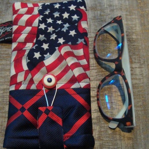 May include: Eyeglass case with an American flag design, featuring red and white stripes and a blue field of stars. The case has a button closure and is paired with eyeglasses with a tortoiseshell pattern frame.
