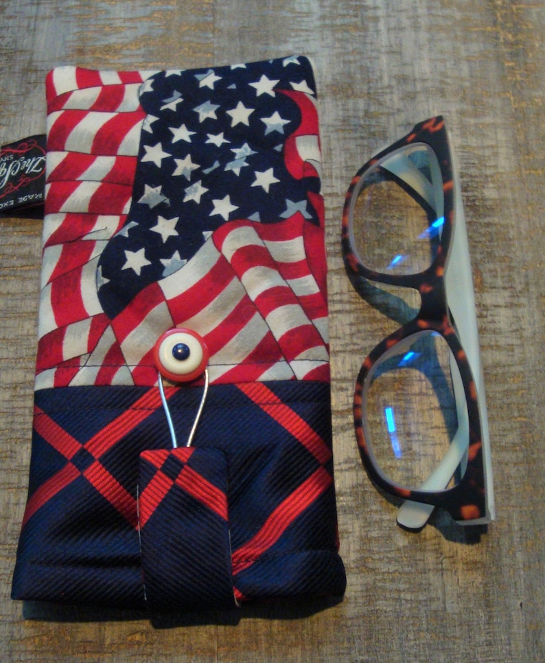 May include: Eyeglass case with an American flag design, featuring red and white stripes and a blue field of stars. The case has a button closure and is paired with eyeglasses with a tortoiseshell pattern frame.