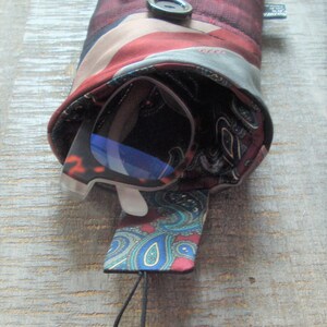May include: A burgundy and patterned glasses case made from repurposed silk ties. The case features a button closure and a paisley interior. A pair of glasses with blue lenses is partially visible inside the case.