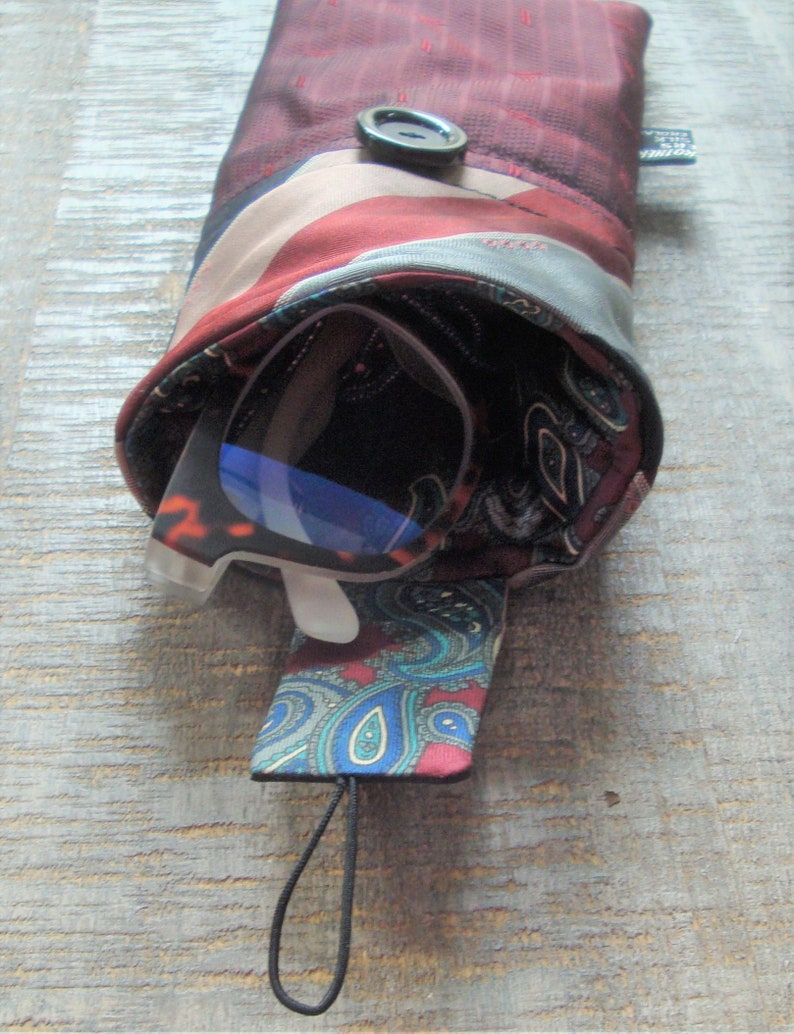 May include: A burgundy and patterned glasses case made from repurposed silk ties. The case features a button closure and a paisley interior. A pair of glasses with blue lenses is partially visible inside the case.