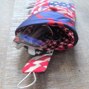 May include: A patriotic glasses case featuring a red, white, and blue American flag design. The case is made of fabric and has a button closure. A pair of sunglasses with a tortoiseshell frame is partially visible inside the case.