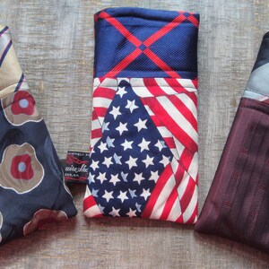 May include: Three fabric eyeglass cases. One case features a navy blue floral pattern with a tan and red striped top. Another has a US flag design. The third case has a burgundy textured pattern with a red and gray striped top.