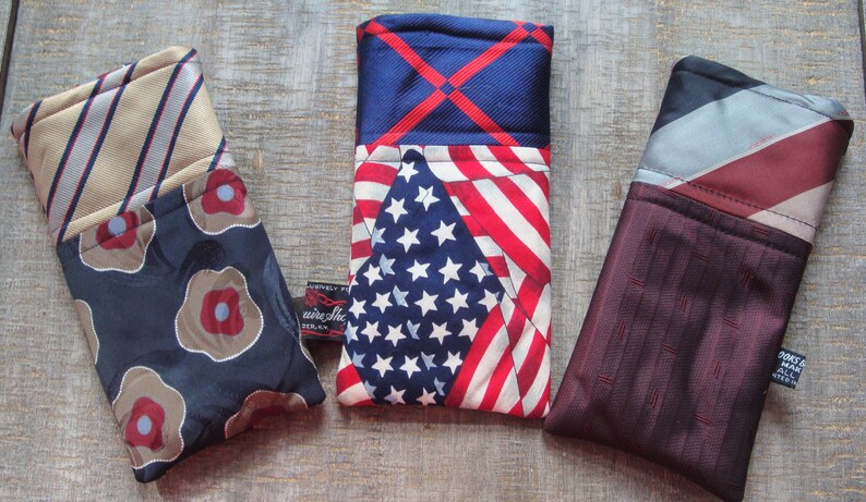 May include: Three fabric eyeglass cases. One case features a navy blue floral pattern with a tan and red striped top. Another has a US flag design. The third case has a burgundy textured pattern with a red and gray striped top.