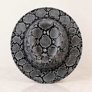 May include: A wide-brimmed fedora hat featuring a high-contrast black and white snakeskin pattern. The hat has a structured crown and a circular brim, with the repeating reptilian scale print covering the entire exterior surface of the headwear.