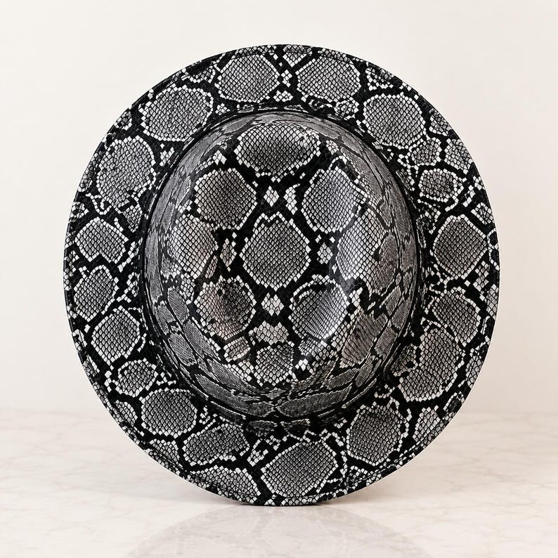 May include: A wide-brimmed fedora hat featuring a high-contrast black and white snakeskin pattern. The hat has a structured crown and a circular brim, with the repeating reptilian scale print covering the entire exterior surface of the headwear.