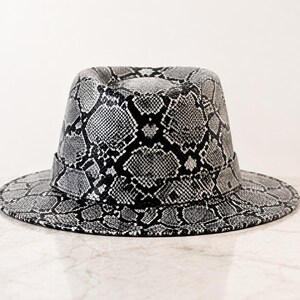 May include: A fedora hat featuring a monochromatic snakeskin pattern in shades of black, white, and gray. The hat has a structured crown with a central crease and a wide, flat brim, finished with a matching snakeskin band around the base.