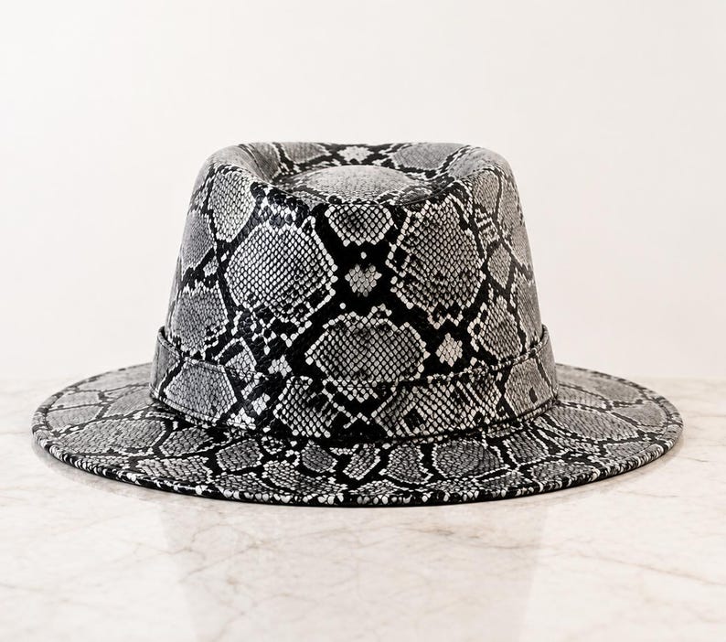 May include: A fedora hat featuring a monochromatic snakeskin pattern in shades of black, white, and gray. The hat has a structured crown with a central crease and a wide, flat brim, finished with a matching snakeskin band around the base.