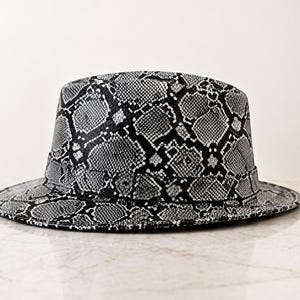 May include: A fedora hat featuring a black and white snakeskin pattern throughout the entire surface. The hat has a structured crown and a flat brim, with a matching band encircling the base of the crown, creating a cohesive and uniform appearance.