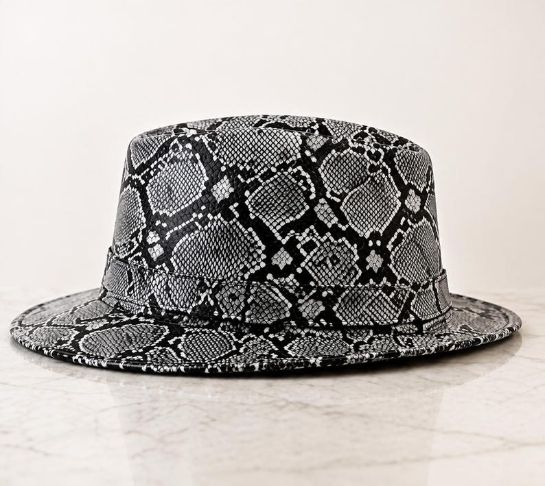 May include: A fedora hat featuring a black and white snakeskin pattern throughout the entire surface. The hat has a structured crown and a flat brim, with a matching band encircling the base of the crown, creating a cohesive and uniform appearance.