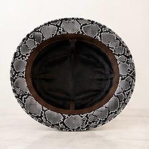 May include: A bucket hat featuring a black and white snakeskin pattern on the exterior brim. The interior of the hat is lined with solid black fabric and includes a brown inner sweatband that circles the crown of the hat.