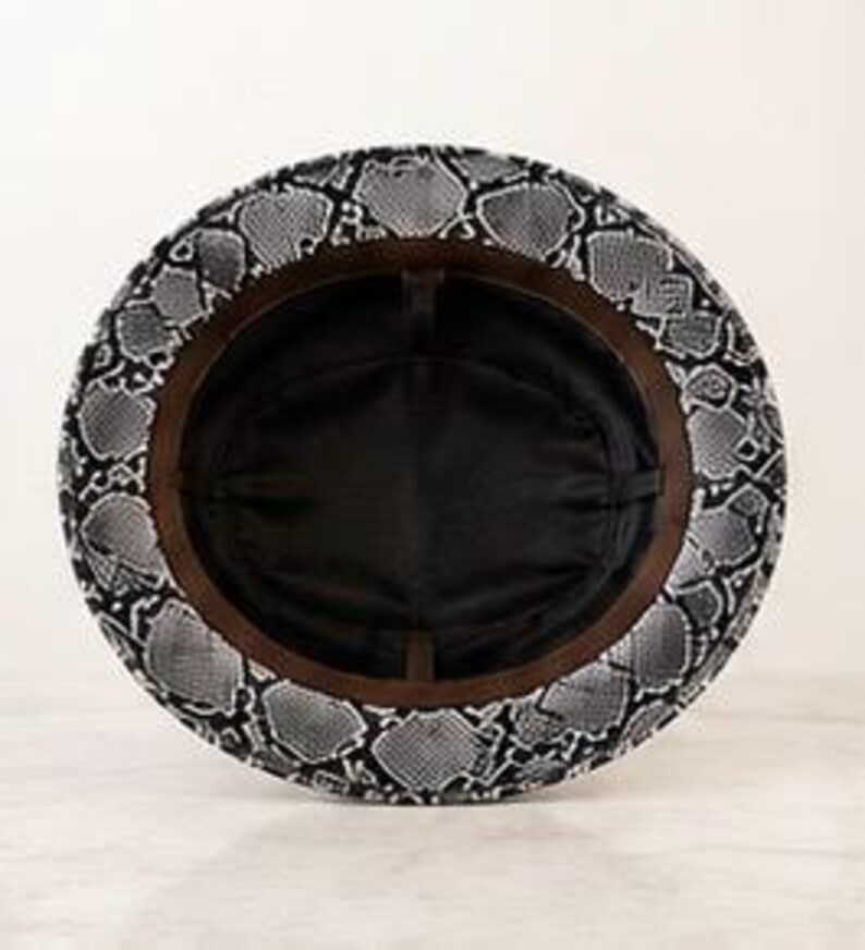 May include: A bucket hat featuring a black and white snakeskin pattern on the exterior brim. The interior of the hat is lined with solid black fabric and includes a brown inner sweatband that circles the crown of the hat.