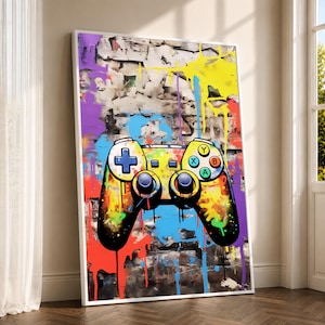 Puede incluir: Una impresión de arte mural vertical enmarcada que presenta una ilustración estilo grafiti vibrante de un mando de videojuegos. El mando se superpone sobre un fondo de pared de ladrillo texturizado con salpicaduras de pintura amarilla, azul, roja y morada.