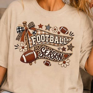 May include: A beige short-sleeve t-shirt featuring a graphic print with the text FOOTBALL SEASON. The design includes a pennant flag, footballs, flowers, stars, and a plaid bow, all rendered in shades of brown, tan, and black with leopard print.