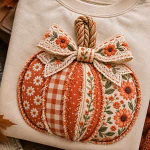 Könnte beinhalten: Ein beiges Sweatshirt mit einer großen, dekorativen Kürbis-Applikation. Der Kürbis besteht aus verschiedenen Stoffbahnen, darunter orangefarbenes Vichy-Karo, weiße Gänseblümchenmuster und florale Designs, gekrönt von einem geflochtenen Seilstiel.