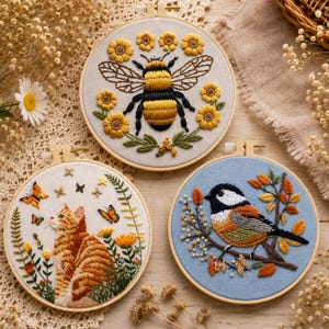 May include: Three circular embroidery hoops featuring detailed needlework. The top hoop shows a bumblebee surrounded by yellow flowers. The bottom left hoop depicts an orange tabby cat with butterflies. The bottom right hoop shows a bird on a branch.