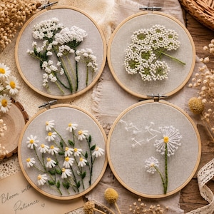 May include: Four circular wooden embroidery hoops containing fabric with hand-stitched floral designs. The top left features white yarrow, the top right shows a white flower cluster, the bottom left displays daisies, and the bottom right depicts dandelions.