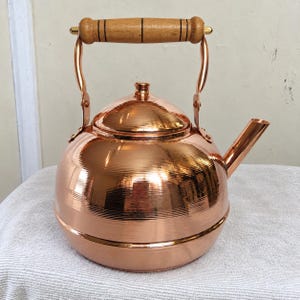 May include: A polished copper tea kettle with a rounded body and a horizontal ribbed texture. It features a removable lid with a small knob, a curved spout, and a hinged handle with a cylindrical light brown wooden grip and black accents.