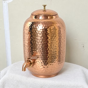 May include: A cylindrical copper beverage dispenser featuring a hammered texture across its exterior surface. The vessel includes a matching copper lid with a small central knob and a functional metal spigot attached to the lower front section.