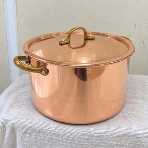 May include: A polished copper stockpot with a matching lid, featuring two curved brass side handles and a single arched brass handle on the lid. The metal surfaces are smooth and reflective, showcasing the warm, metallic orange-brown hue of copper.