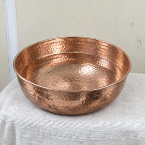 May include: A large, round, hammered copper basin with a polished metallic finish. The bowl features a distinct dimpled texture across its entire surface, creating a reflective appearance. The rim is smooth and rounded, forming a wide, open vessel.