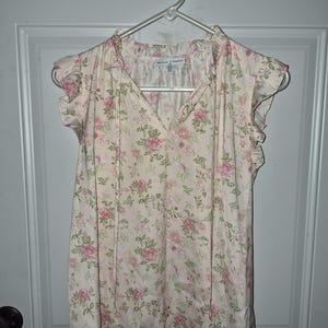 May include: A cream-colored sleeveless blouse featuring a pink and green floral pattern, a V-neckline with thin tie strings, and ruffled cap sleeves. The interior label reads Maison d'Amélie. The garment is displayed on a white plastic hanger.