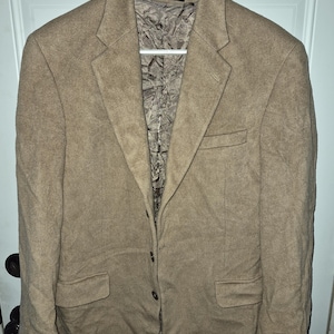 Könnte beinhalten: Ein beigefarbener Blazer mit strukturierter Oberfläche, ausgestattet mit steigendem Revers, drei Knöpfen auf der Vorderseite und zwei Pattentaschen. Das Innere ist mit braunem, knittrigem Stoff gefüttert. Ein Etikett am Kragen zeigt Made in Fiji Islands, dry clean only.