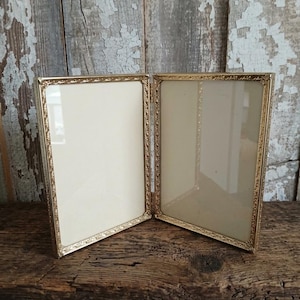May include: A double-hinged picture frame featuring two rectangular panels with a metallic gold finish. Each frame border displays a delicate, repeating embossed floral pattern. The structure is designed to stand upright, holding two clear glass panes.
