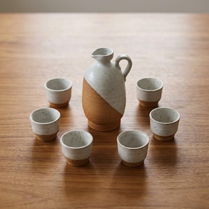 May include: A ceramic sake set consisting of one pitcher with a handle and five small cups. Each piece features a two-tone design with a speckled white glaze on the upper portion and a natural, unglazed terracotta-coloured base at the bottom.