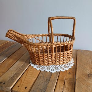 May include: A light brown woven wicker basket featuring an elongated, cradle-like shape with an open top, a single arched handle positioned on one side, and a pointed, extended end, crafted from natural reed material with a visible weave pattern.