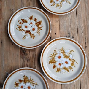 Peut inclure: Quatre assiettes rondes en céramique avec une base couleur crème et un bord marron. Chaque assiette est ornée d'un motif floral central composé de fleurs blanches aux centres orange entourées de feuilles et de tiges marron en bouquet.
