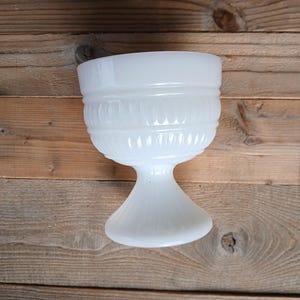 Peut inclure: Un gobelet en verre opalin blanc présentant un bord supérieur lisse, une bande centrale avec des crêtes verticales en relief et une base sur pied avec un motif cannelé. Le verre a un aspect opaque et laiteux avec une finition brillante.