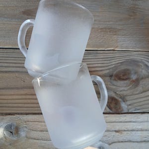 Peut inclure: Deux tasses en verre transparent avec une finition givrée et mate. Chaque tasse présente une anse lisse et arrondie ainsi qu'un corps cylindrique. Les tasses sont empilées en angle, mettant en valeur la surface translucide et texturée.