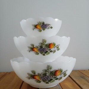Peut inclure: Un ensemble de trois bols gigognes en verre opalin blanc avec des bords festonnés. Chaque bol présente un motif de fruits coloré représentant des poires orange, des feuilles vertes et des grappes de raisins violets sur une branche.