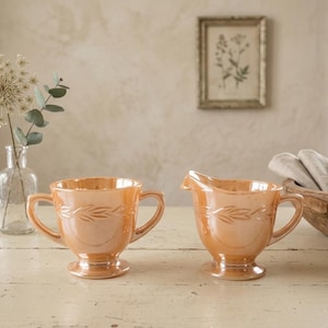 Peut inclure: Une paire de récipients en verre irisé couleur pêche, composée d'un sucrier à deux anses et d'un pot à lait assorti. Les deux pièces présentent une base sur pied et un motif de feuilles en relief entourant la partie supérieure.