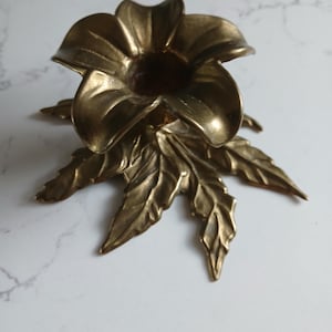 May include: A brass candle holder shaped like a flower with five prominent petals forming the cup and several textured leaves extending outwards at the base. The metal has a warm, metallic gold tone with a polished finish and visible contours.