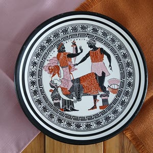 Peut inclure: Une assiette décorative ronde ornée d'une illustration de style grec en noir et blanc représentant des personnages en toge, dont une personne allongée et une autre debout tenant une épée. La bordure présente un motif géométrique grec.