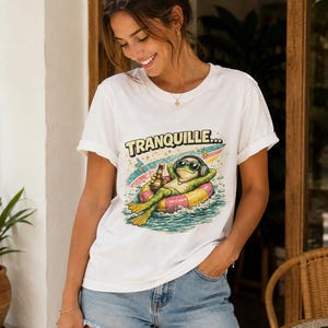 Peut inclure: Un t-shirt blanc à manches courtes avec une illustration d'une grenouille verte portant des lunettes de soleil et un casque, flottant sur une bouée colorée dans l'eau avec une bouteille et le texte TRANQUILLE au-dessus.