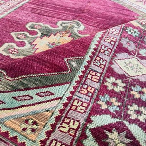 May include: A magenta area rug featuring a geometric pattern with accents in light blue, cream, and dark gray. The rug displays a central medallion design and a detailed border with repeating floral motifs, showcasing a textured pile surface.