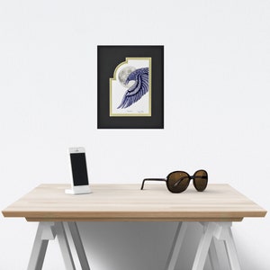 May include: Framed artwork featuring a blue and white illustration of a bird's wing in front of a full moon. The artwork is in a black frame with a gold border. A white smartphone and black sunglasses rest on a light wood table.