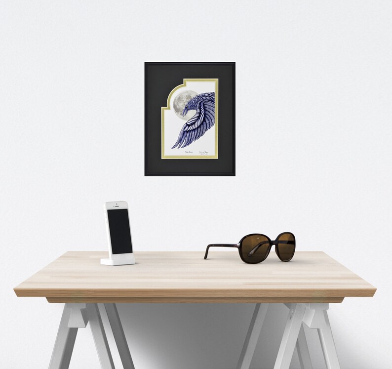 May include: Framed artwork featuring a blue and white illustration of a bird's wing in front of a full moon. The artwork is in a black frame with a gold border. A white smartphone and black sunglasses rest on a light wood table.