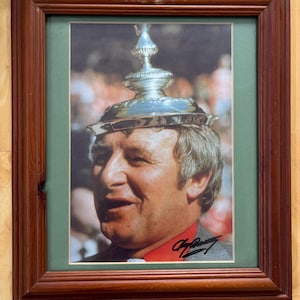 May include: A framed photograph of a person wearing a silver trophy as a hat. The person has light hair and is wearing a red shirt under a gray jacket. The photograph features a black signature at the bottom and is encased in a dark wood frame.