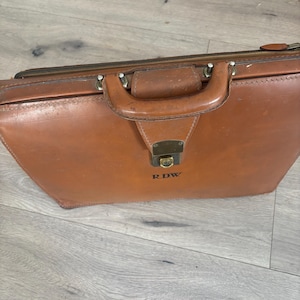 May include: A tan leather briefcase featuring a single top handle, a metal locking clasp, and a zipper closure along the top edge. The front of the briefcase is embossed with the letters R.D.W. in a dark, capitalized font centered below the clasp.