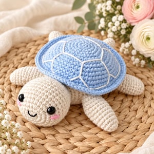 May include: A crocheted turtle toy featuring a light blue shell with white stitched detailing and a cream-colored body. The turtle has two black plastic eyes, a small embroidered black mouth, and pink stitched blush marks on its cheeks.
