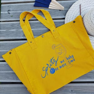 Peut inclure: Un sac fourre-tout en toile jaune vif orné d'un motif bleu représentant un verre à cocktail avec une tranche d'agrume et l'inscription "Spritz me baby one more time" en bleu. Le sac possède deux anses et des boutons-pression métalliques.