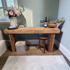 May include: A rectangular wooden console table with a natural finish and thick block legs. The surface features the text HandmadeFurnituree. It is styled with a white vase of flowers, a wine glass, a bottle of wine, and a wooden bowl with a pineapple.