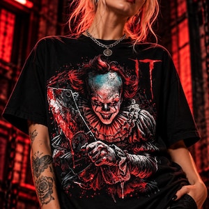 Puede incluir: Una camiseta negra de manga corta con un estampado gráfico del personaje Pennywise de la película IT. El payaso aparece con pelo rojo, un cuello con volantes y un gran cuchillo de carnicero manchado de sangre. La palabra IT aparece en rojo.