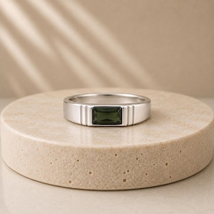May include: A silver-toned band ring featuring a rectangular, dark olive-green gemstone set horizontally in the center. The metal band has a smooth, polished finish with two vertical grooves flanking each side of the central stone setting.